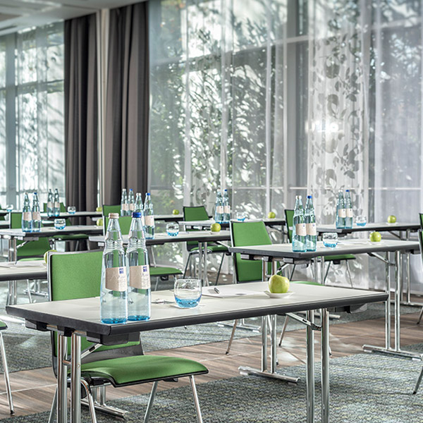 Heller Konferenzraum mit grünen Stühlen, Tischen voller Wasserflaschen, Äpfeln und Notizblöcken