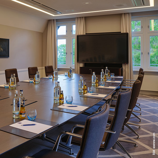 Konferenzraum mit großem Tisch, Stühlen, Getränken und Schreibmaterialien vor einem Bildschirm