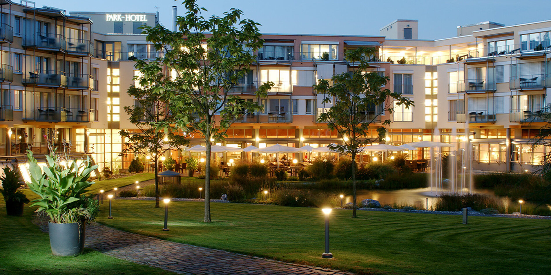 Abendliche Ansicht des Park-Hotels mit beleuchteter Fassade, gepflegtem Garten, Teich und Terrasse