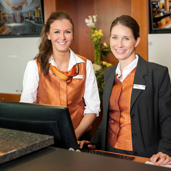 Zwei Frauen in orangefarbenen Uniformen stehen lächelnd hinter einem Empfangstresen
