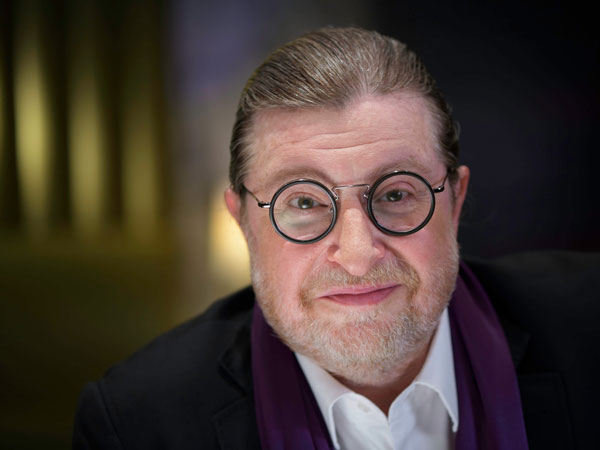 Mann mit zurückgekämmten Haaren, weißem Hemd und dunklem Sakko mit violettem Schal